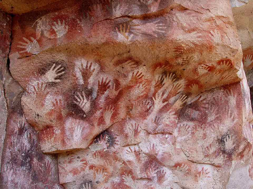 Hundreds of stencilled hands in red, white and ochre on a rock wall.
